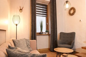 Een woonkamer met een bank, fauteuil, rond houten tafeltje, vloerlamp en een raam met gordijnen.