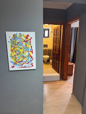 Un pasillo con una puerta de madera que da a un baño, un cuadro abstracto y colorido en la pared, y un suelo de baldosas.