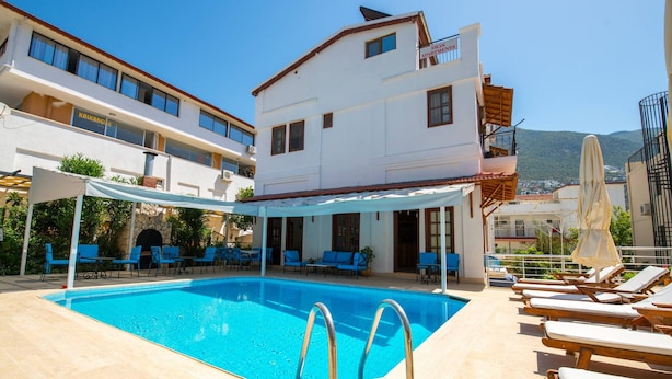 Una piscina con sillones y sombrillas, rodeada de edificios con balcones y un cielo despejado.