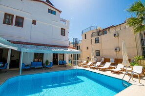 Una piscina con sillas de descanso y un área de estar cubierta, rodeada de edificios con balcones.