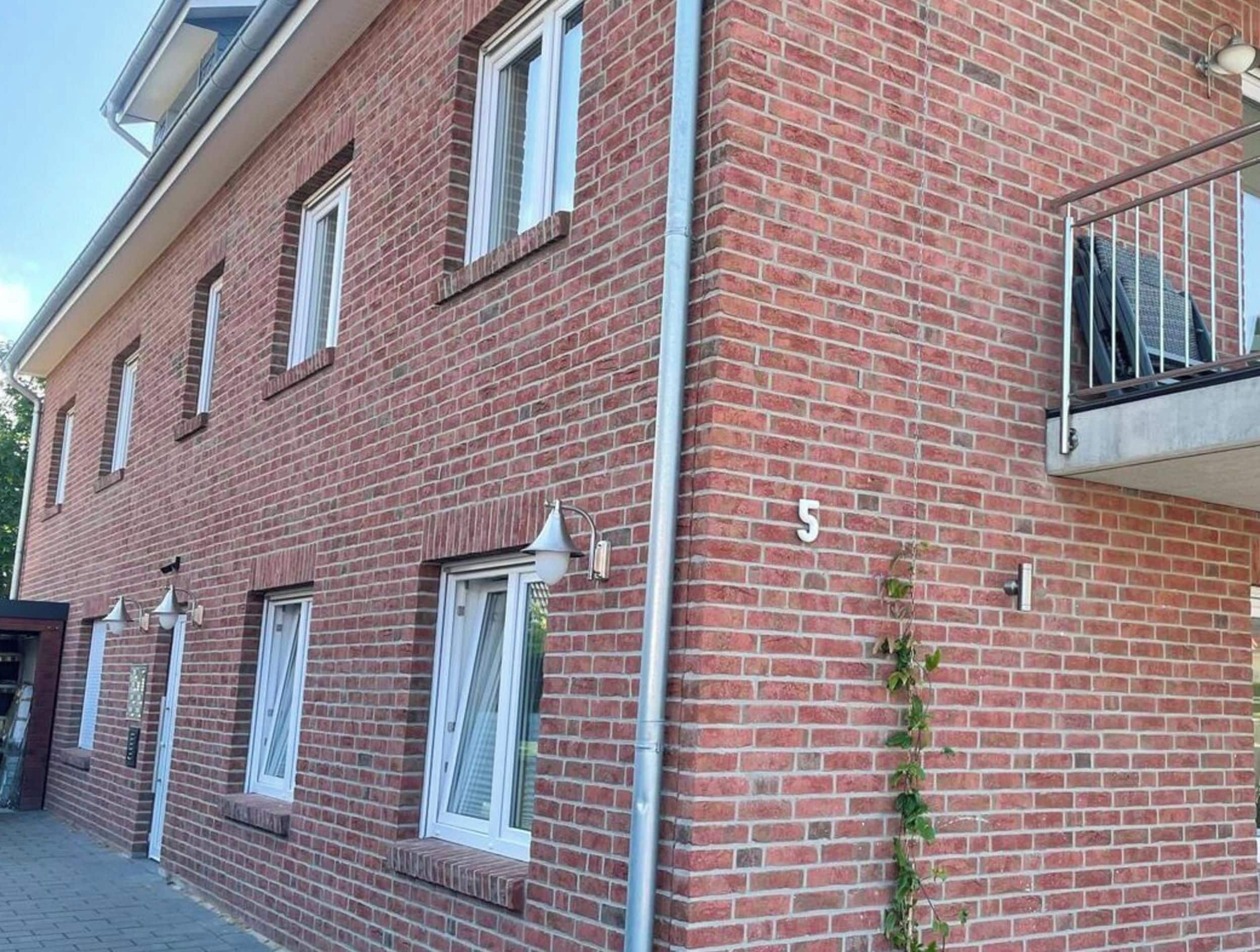 Un edificio de ladrillo rojo con ventanas de marco blanco, un número '5' visible en la fachada y un pequeño balcón con barandilla de metal.