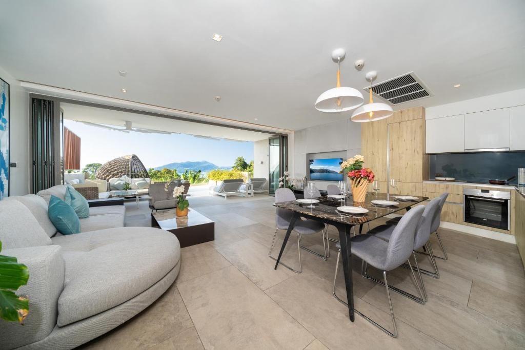 A modern living room with a sofa, dining table, and chairs. There is a kitchen area with a TV and a large window overlooking a mountainous landscape.