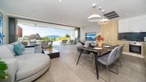 Een moderne woonkamer met een bank, eettafel en stoelen. Er is een keukenhoek met een televisie en een groot raam met uitzicht op een bergachtig landschap.