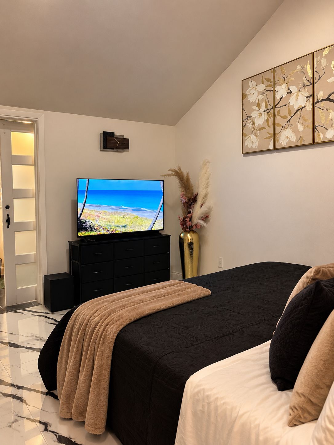 Ein Schlafzimmer mit einem Bett, einem Fernseher auf einer Kommode, einer Vase mit Trockenblumen und einem Gemälde an der Wand.