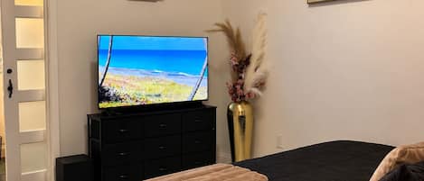 A bedroom with a bed, a television on a dresser, a vase with dried flowers, and a painting on the wall.