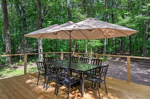 Eine Holzterrasse mit Tisch und Stühlen unter einem großen Sonnenschirm, umgeben von einem Wald.