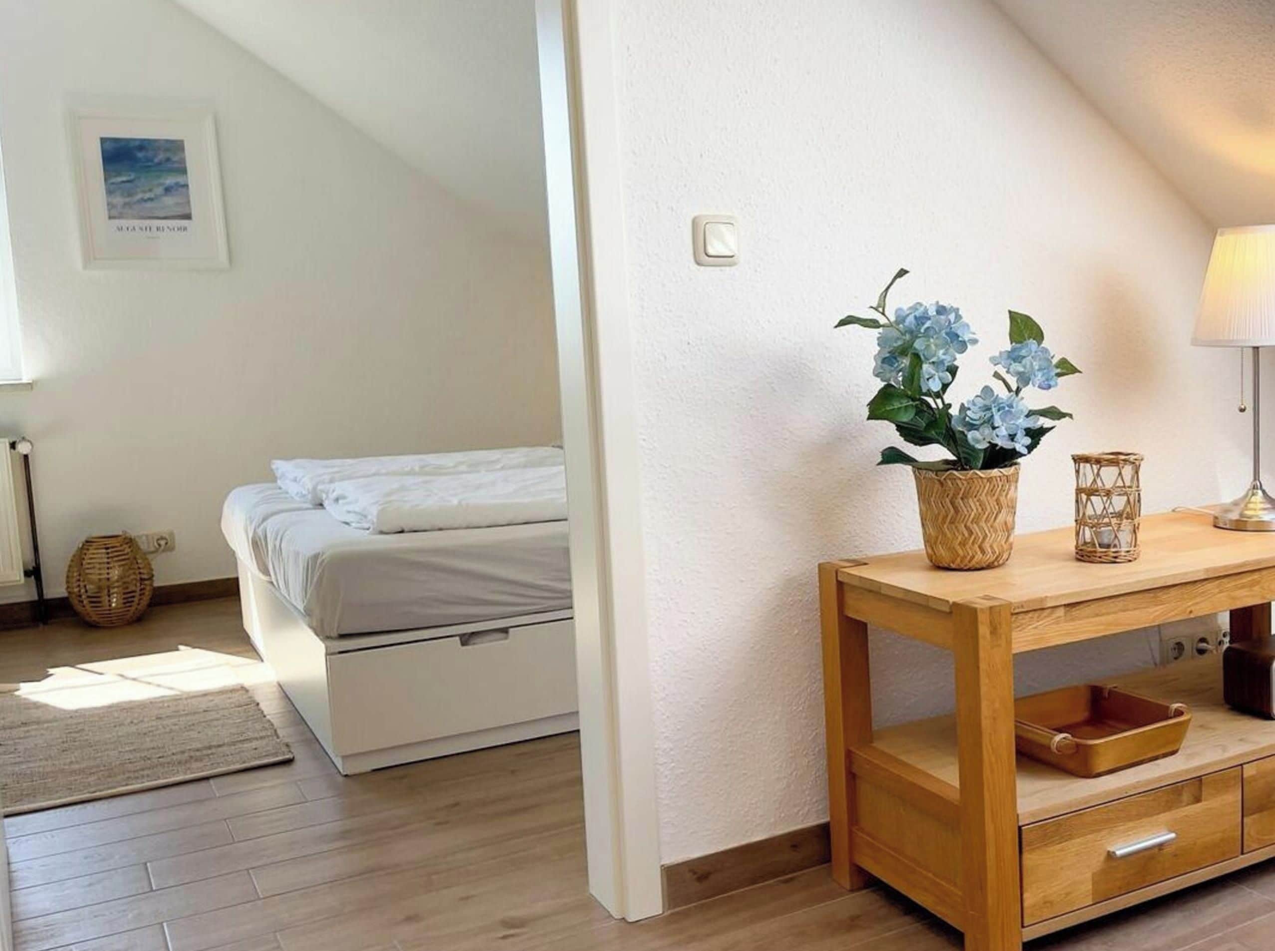 Un dormitorio con cama, una mesita de noche con lámpara, una cesta de mimbre y un cuadro enmarcado en la pared.