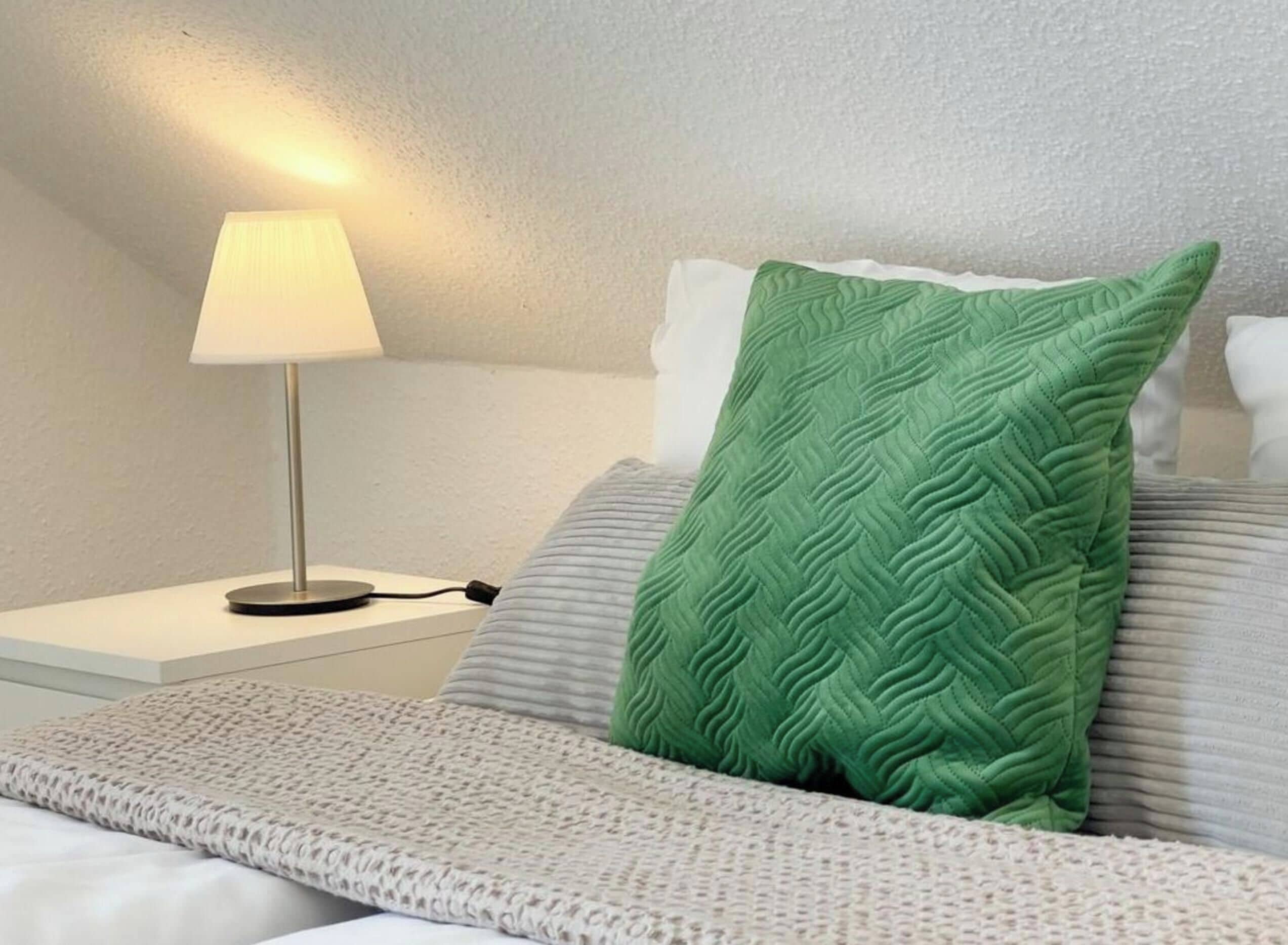 En pænt redt seng med en grøn pude med struktur, en hvid pude og en grå pude. Der er et hvidt natbord med en lampe og et strømkabel.