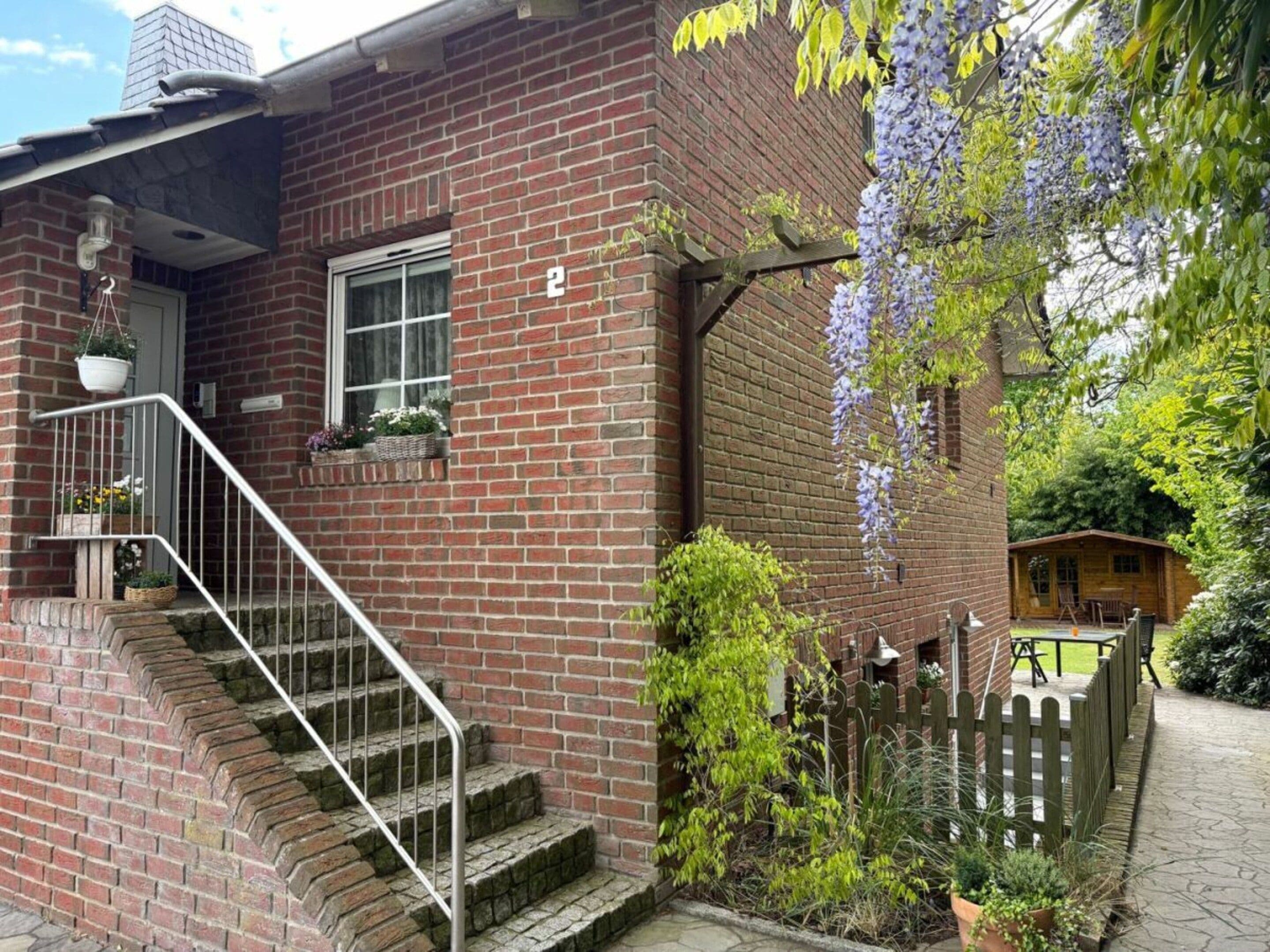 Une maison en briques avec un escalier, une clôture en bois et un jardin où poussent des fleurs violettes.
