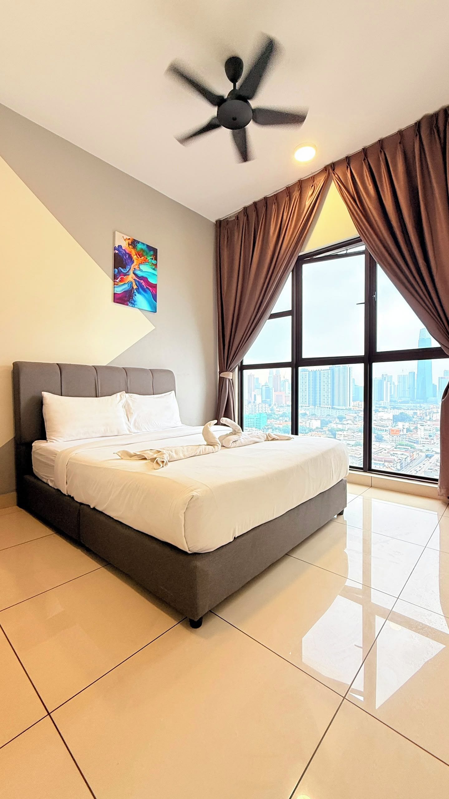 A modern hotel room with a large bed, a ceiling fan, a city view through the window, and a painting on the wall.