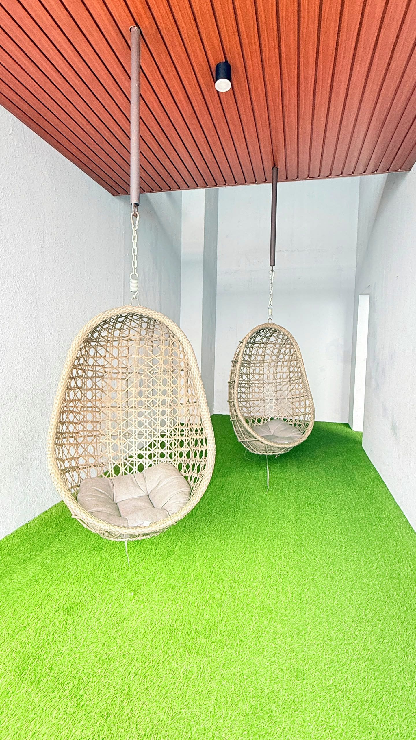Two hanging wicker chairs with cushions on a green artificial turf floor, suspended from a wooden ceiling with visible beams.