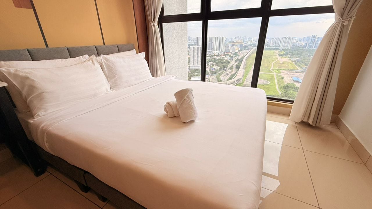 Una cama bien hecha con sábanas blancas y una toalla, una ventana con cortinas, y vista a un paisaje urbano.