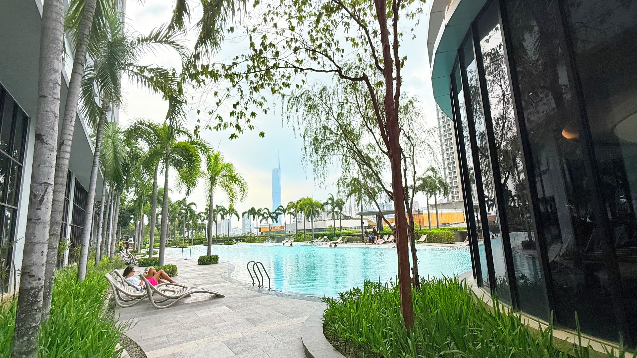 Zona de piscina con sillones reclinables, palmeras y un edificio moderno al fondo.