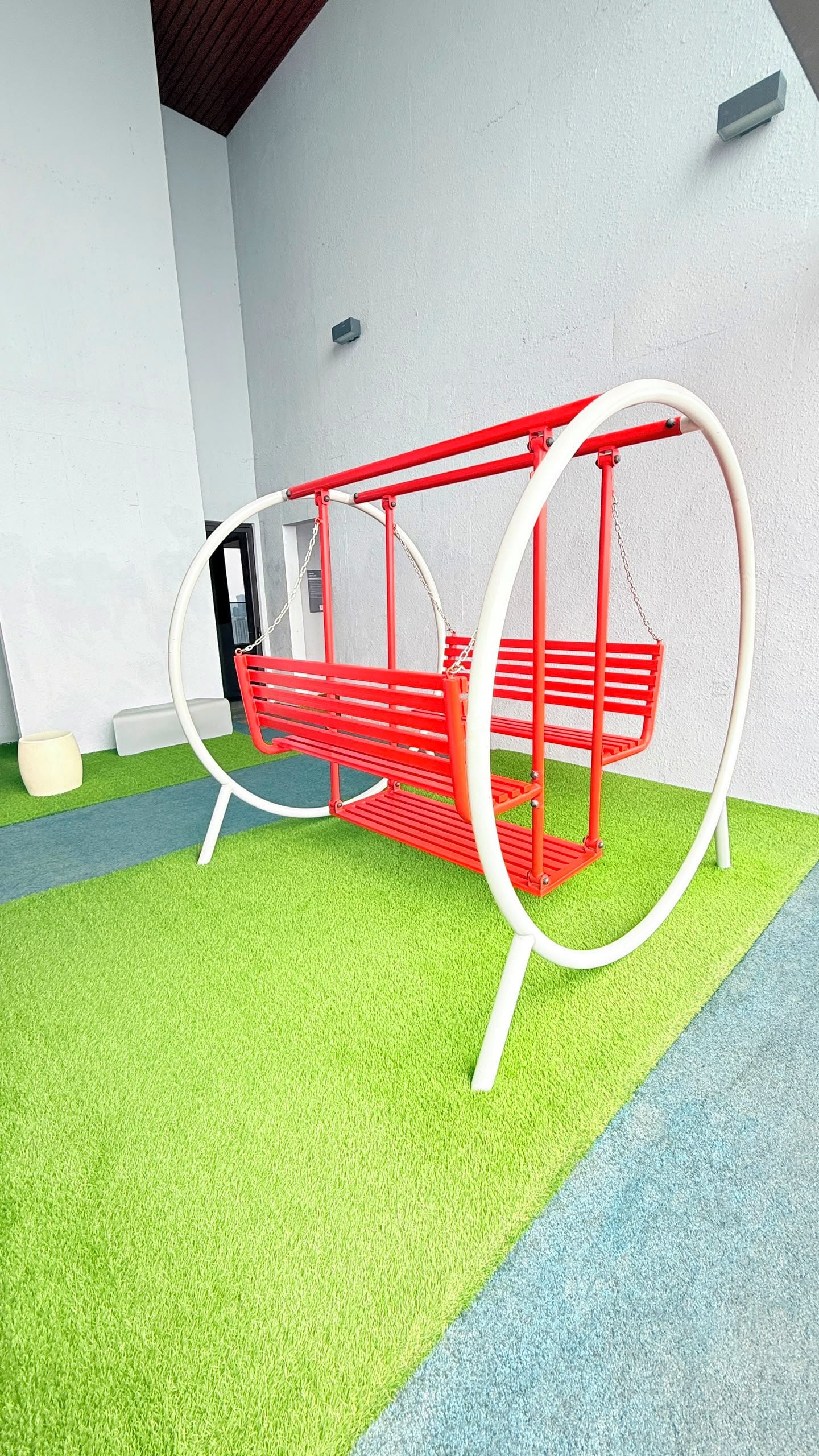 A red swing set with white supports and chains, located on a green artificial turf area.