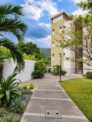 A pathway leading to a building with a modern design, surrounded by lush greenery and a clear sky. - Serene Smart Suite ● Devon House Ice-Cream ● Gated ● Smart Stay (Kingston)