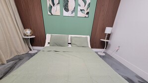 Un dormitorio con cama, dos almohadas, pared verde, panel de madera, una mesita redonda con lámpara y cortina.