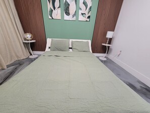 Makuuhuone, jossa on sänky, kaksi tyynyä, vihreä seinä, puupanelointi, pieni pyöreä pöytä, jossa on lamppu, ja verho.