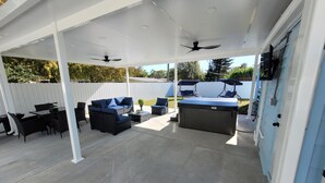 Un patio exterior con un área de estar cubierta, que incluye un sofá, sillas, una mesa y un jacuzzi.