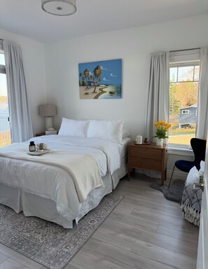 A bedroom with a bed, a nightstand, a lamp, a painting, a window with curtains, and a view of the outdoors.