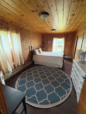 Una habitación de madera con una cama, un aparador, un tapete redondo y una ventana con cortinas.