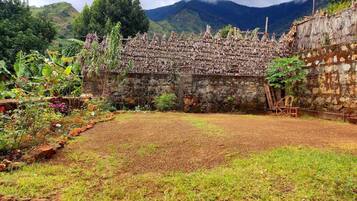 A rustic stone structure with a thatched roof, surrounded by a garden with various plants and a clear view of mountains in the background.