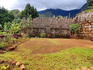 A rustic stone structure with a thatched roof, surrounded by a garden with various plants and a clear view of mountains in the background. - Olympos Camping (Morogoro)