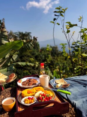 A meal tray with toast, fruit, and a mug, set against a backdrop of lush greenery and a mountain view. - Olympos Camping (Morogoro)