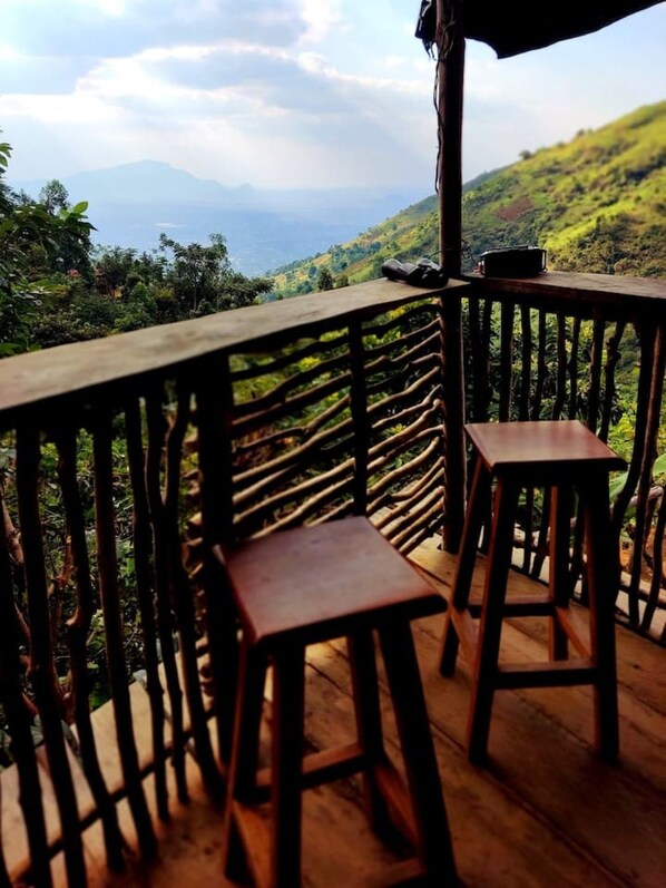 A wooden deck with two stools and a table, overlooking a lush green landscape with hills and mountains in the distance.