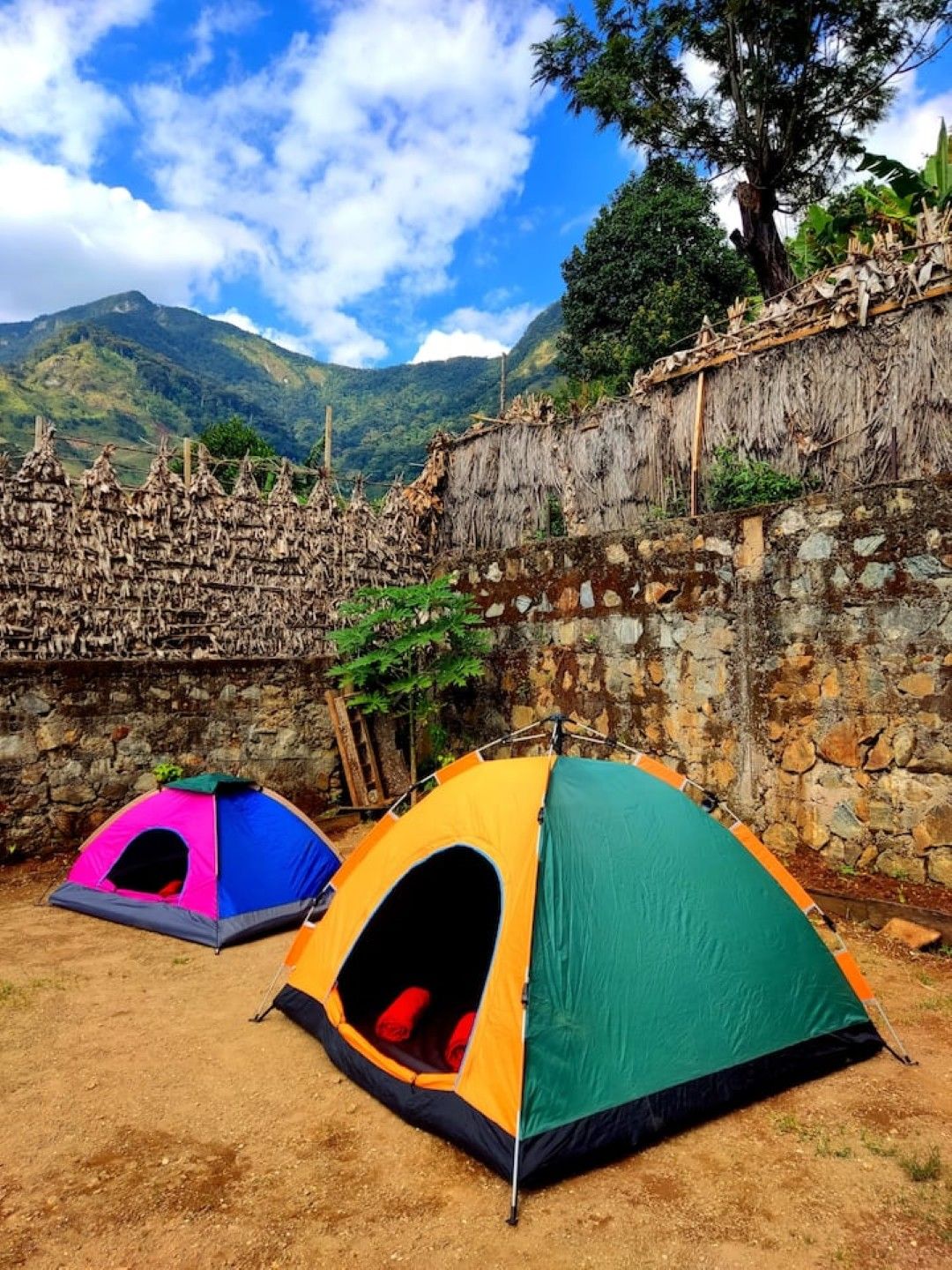 Un campamento con tres tiendas de campaña montadas sobre terreno de tierra, rodeado por un muro de piedra y un paisaje de montañas.