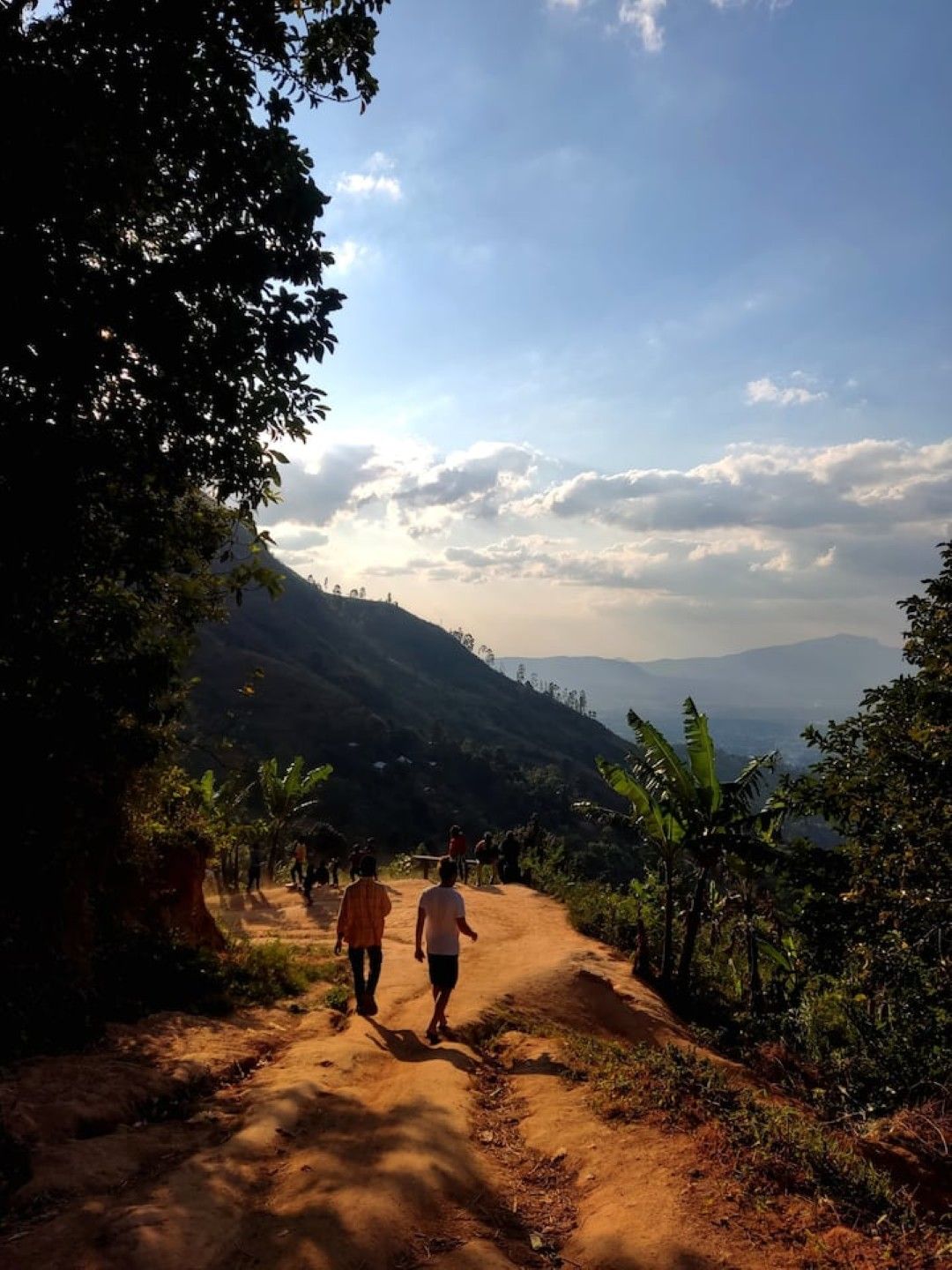 A dirt path with people walking, surrounded by greenery and hills in the background.