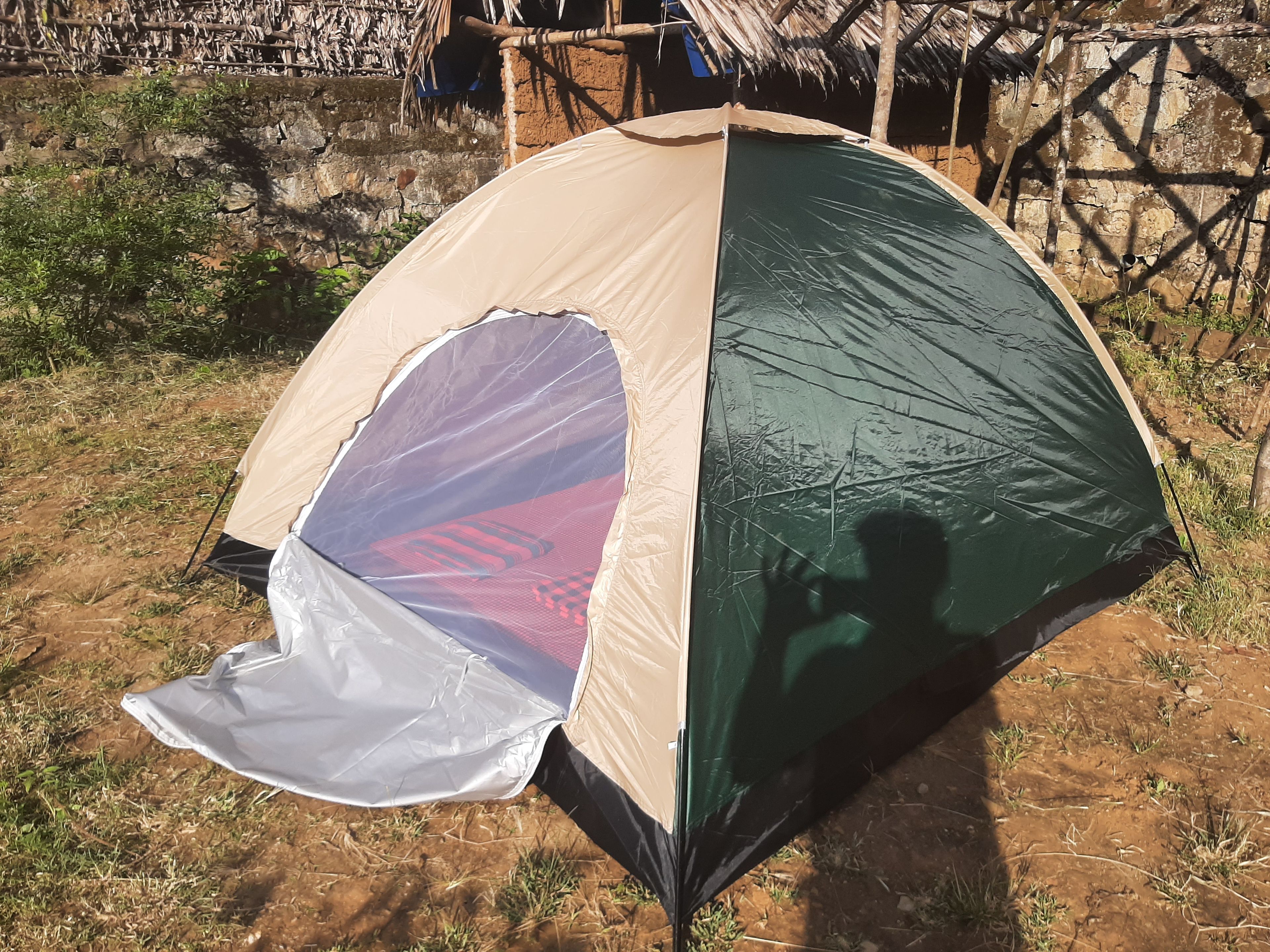 Een koepelvormige tent met een gedeeltelijke zeilafdichting, opgetrokken op een zandgrond met schaarse begroeiing en een rieten constructie op de achtergrond.