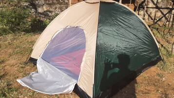 Een koepelvormige tent met een gedeeltelijke zeilafdichting, opgetrokken op een zandgrond met schaarse begroeiing en een rieten constructie op de achtergrond.