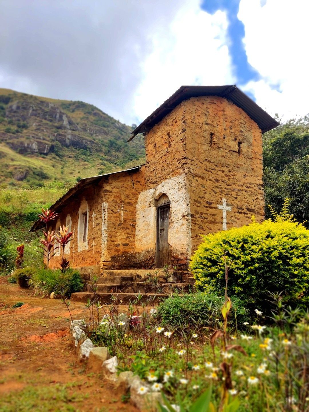 Una iglesia de piedra rústica con un campanario, rodeada de abundante vegetación y una colina al fondo.