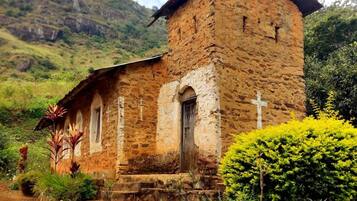Una iglesia de piedra rústica con un campanario, rodeada de abundante vegetación y una colina al fondo.