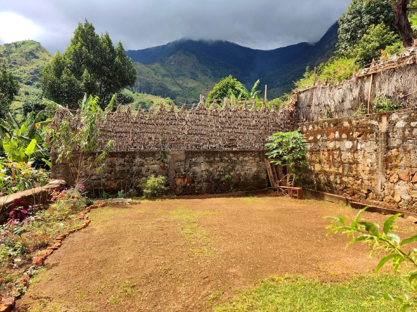 A courtyard with a stone wall, a thatched roof structure, and a view of mountains in the background.