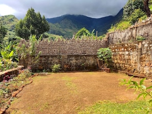 A courtyard with a stone wall, a thatched roof structure, and a view of mountains in the background.