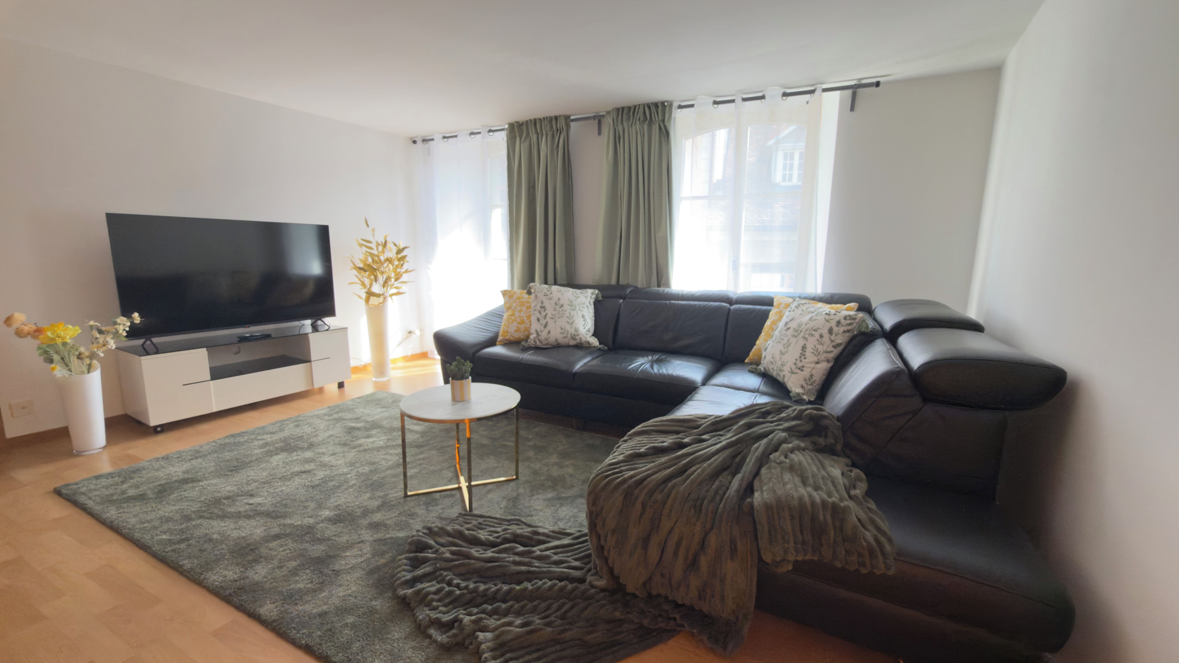 A modern living room with a black leather sofa, a flat-screen TV, a small round coffee table, and a grey rug.