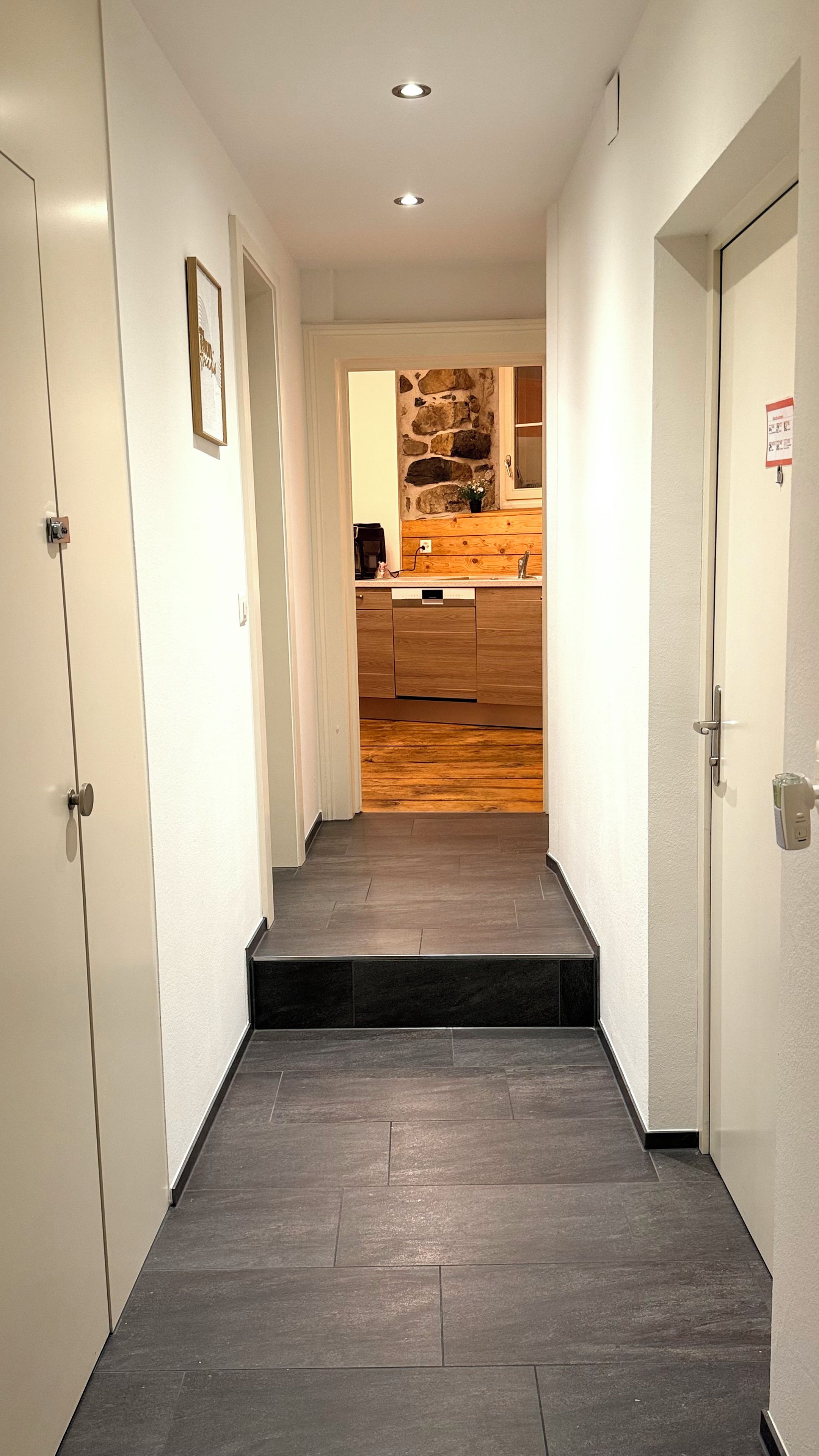 A hallway with a wooden floor leading to a kitchen area with a stone wall backsplash.