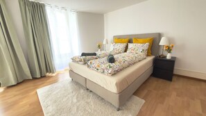 Une chambre à coucher avec un lit, des tables de chevet, une commode, une fenêtre avec des rideaux et un plancher en bois.