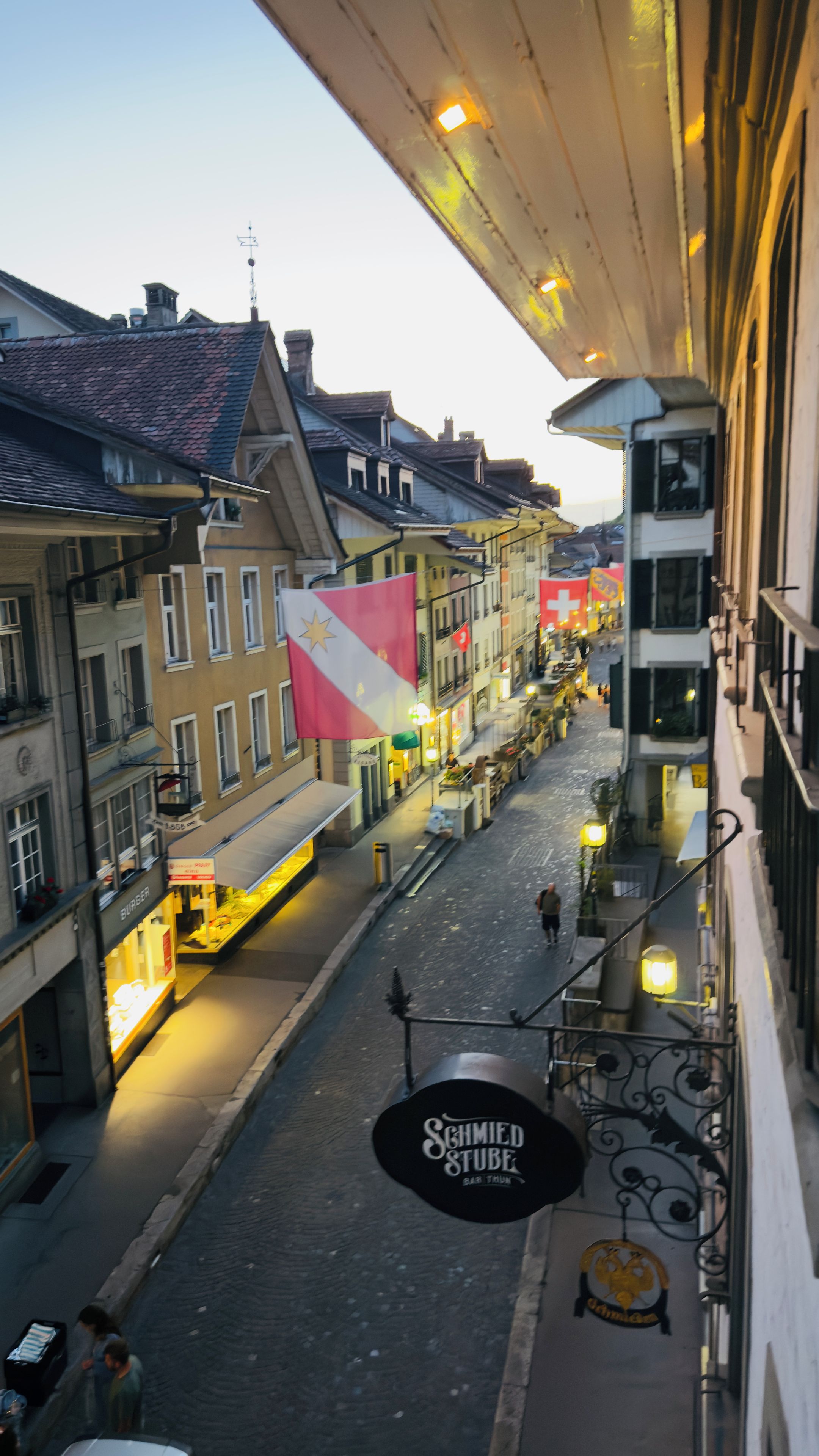 Une vue de rue avec des bâtiments, un drapeau et une enseigne indiquant « Schmied Stube Bar Thun ».
