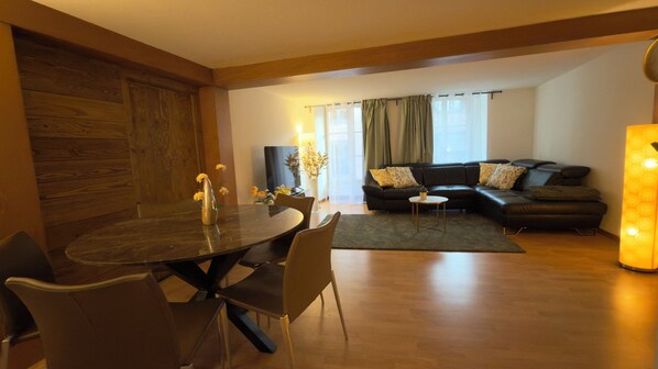 A modern living room with a dining area, featuring a wooden floor, a black leather sofa, and a round dining table with chairs.