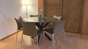 Mesa redonda com tampo de vidro, rodeada por quatro cadeiras, um abajur e uma porta de madeira ao fundo.