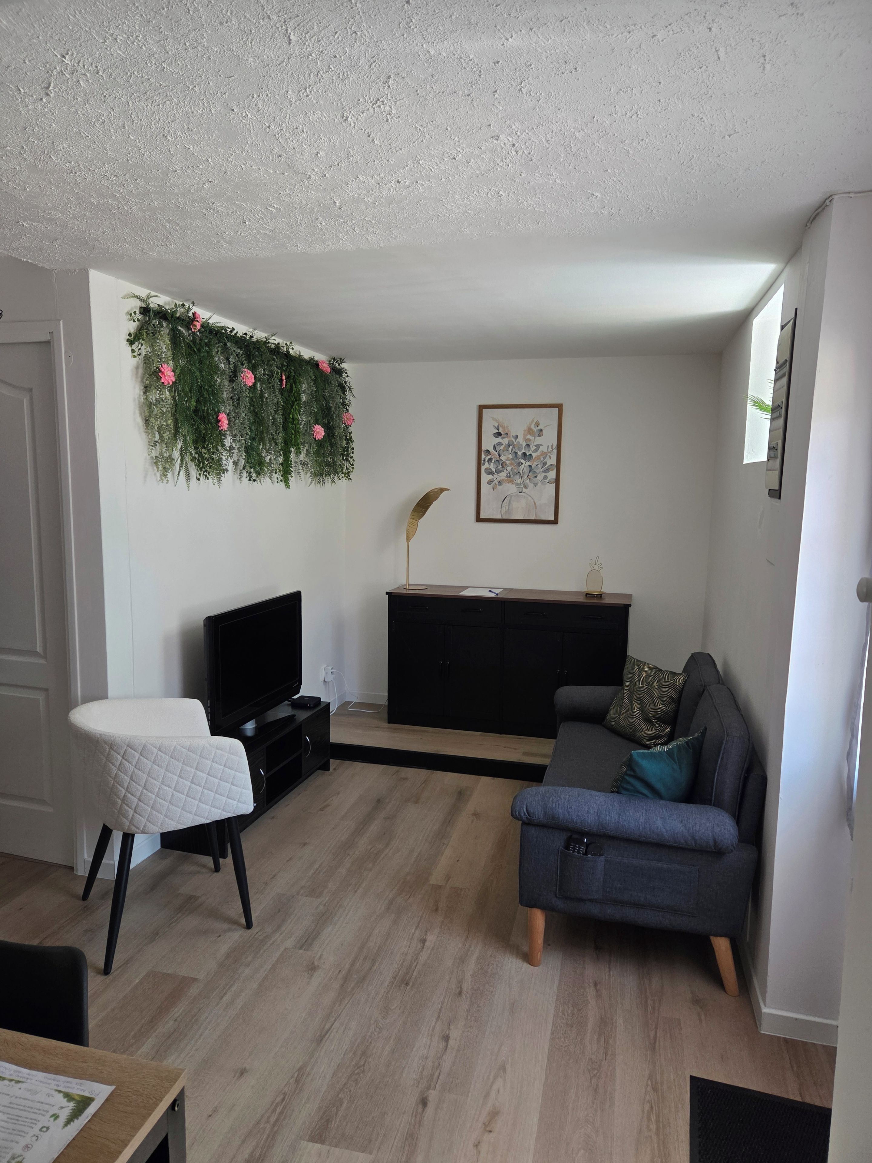A living room with a flat-screen TV, a grey sofa, a white chair, a black entertainment unit, and a decorative plant on the wall.