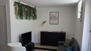 En stue med et fladskærms-tv, en grå sofa, en hvid stol, et sort tv-møbel og en dekorativ plante på væggen.