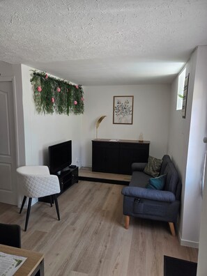 Een woonkamer met een flatscreen televisie, een grijze bank, een witte fauteuil, een zwarte tv-kast en een decoratieve plant aan de muur.
