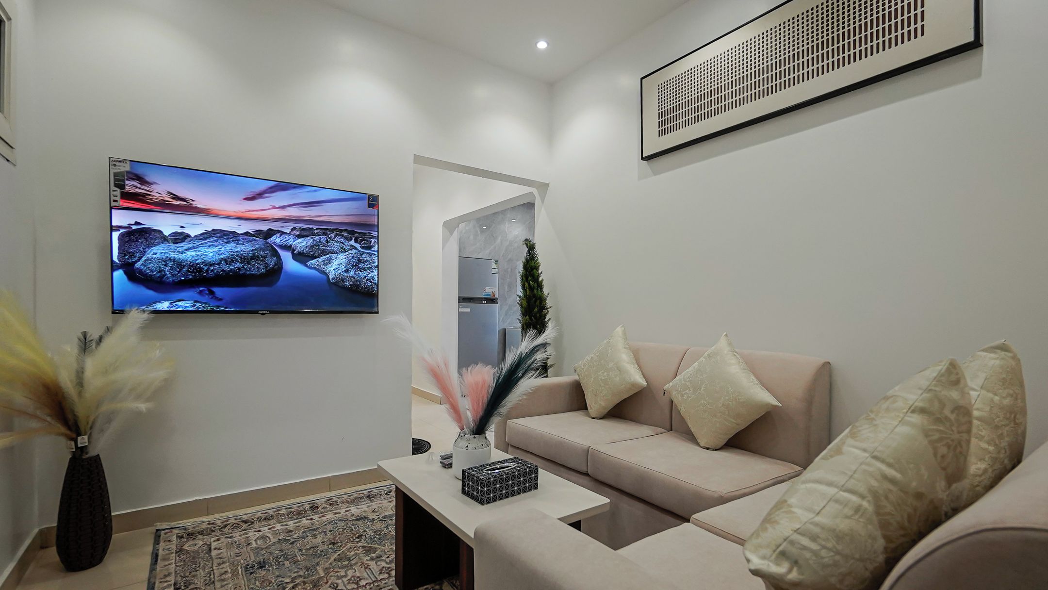 Una sala moderna con un televisor de pantalla plana montado en la pared, un sofá color beige con almohadones decorativos, una mesita con un florero y una caja, y una alfombra estampada en el suelo.