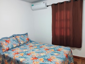 A single bed with a floral bedspread, a brown curtain, and a wall-mounted air conditioning unit. - Cozy studio near Iguazu Falls and the Argentinian border. (Foz do Iguaçu)