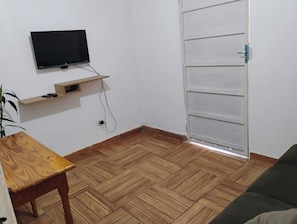 A room with a television mounted on the wall, a wooden floor, a sofa, and a door with a handle. - Cozy studio near Iguazu Falls and the Argentinian border. (Foz do Iguaçu)