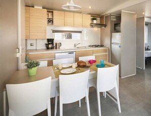 A compact kitchen with wooden cabinets, a white countertop, and a dining area with white chairs. - Sunlit Park Umag Escape (Brtonigla)