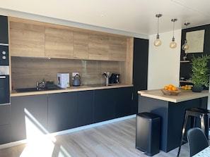 A modern kitchen with wooden cabinets, a black countertop, a kettle, a coffee maker, and a bowl of oranges.